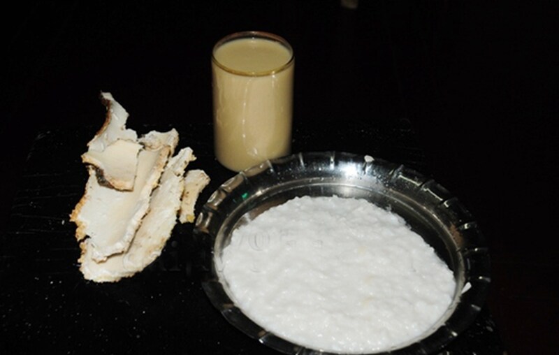 A glass of pale Aati Kashaya beside a plate of white rice porridge and pieces of medicinal bark
