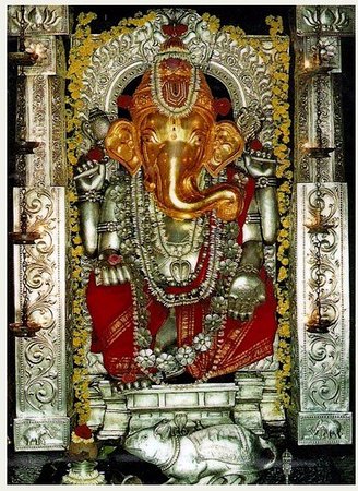 The golden idol of Sri Anegudde Vinayaka adorned with silver ornaments, flower garlands and red cloth, framed by an ornate silver prabhavali in the sanctum