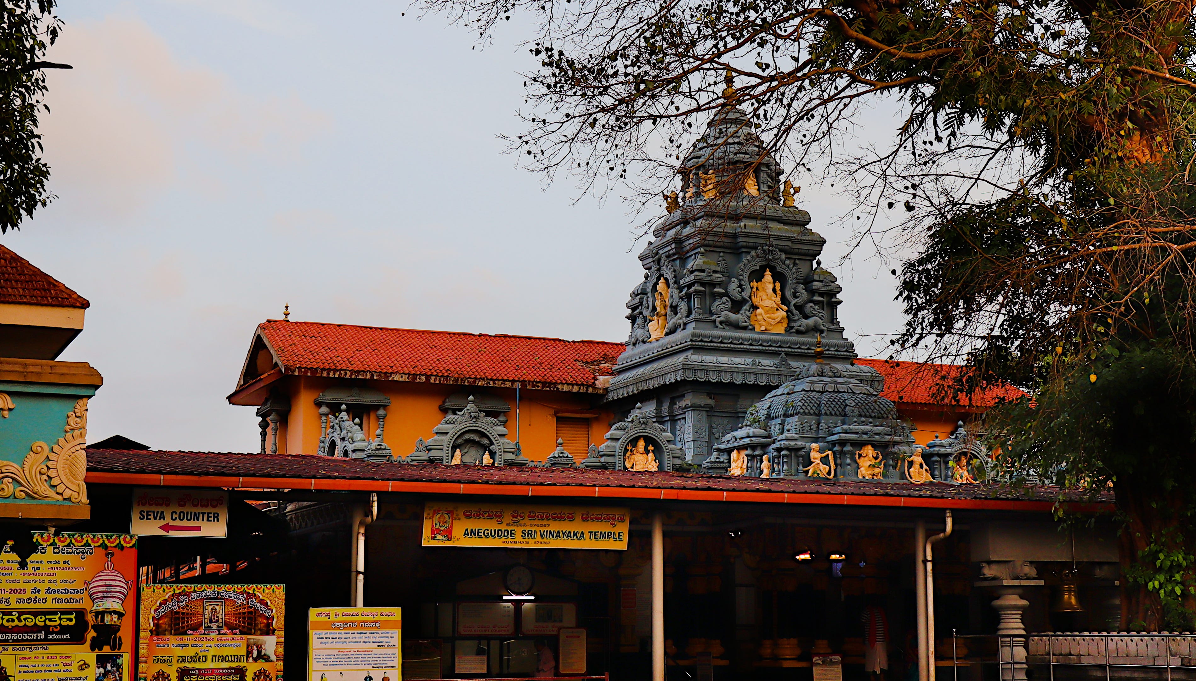 The grey stone vimana of the Anegudde Vinayaka temple rising above orange walls and tiled roofs at dusk, Kumbashi