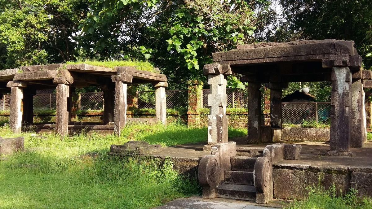 Weathered stone mandapa pavilions with spiral-carved steps standing in a green clearing among trees at Barkur