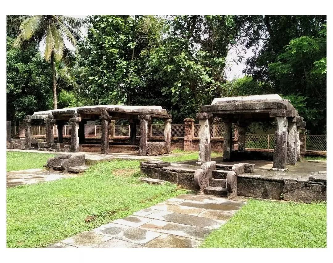 Stone pavilions beside a paved slab pathway leading through a green clearing at Barkur