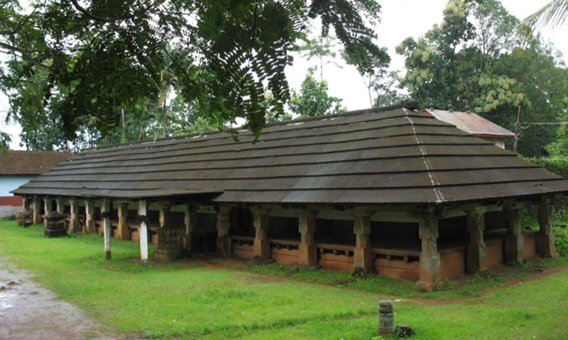 A long tiled-roof temple hall on stone pillars surrounded by greenery at Barkur