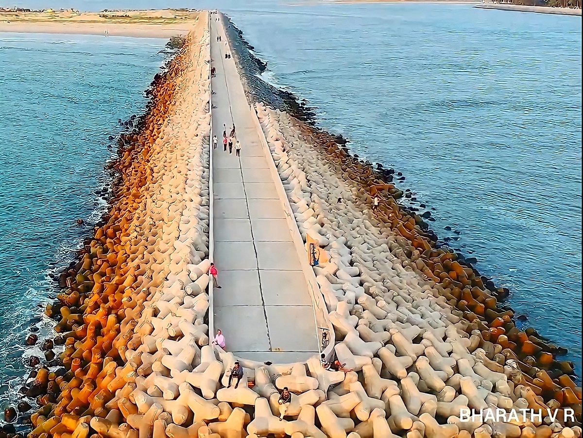 The long tetrapod-and-concrete sea walk breakwater at Gangolli reaching out between river and sea at golden hour, with people strolling along it