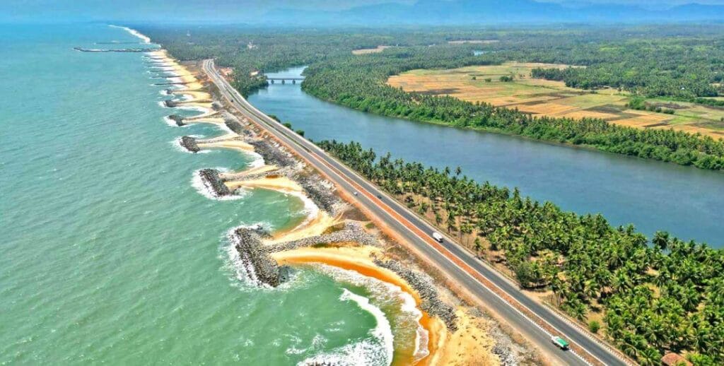 Aerial view of the Maravanthe coast with turquoise sea, rock groynes, the highway and the Souparnika river running parallel inland past paddy fields