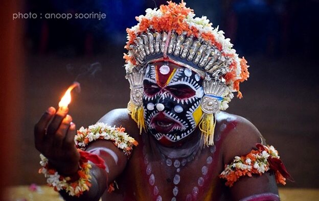 A Bhoota Kola performer with intricate red, white and black facial designs and a silver-and-flower headdress, holding a small lit candle in the dark