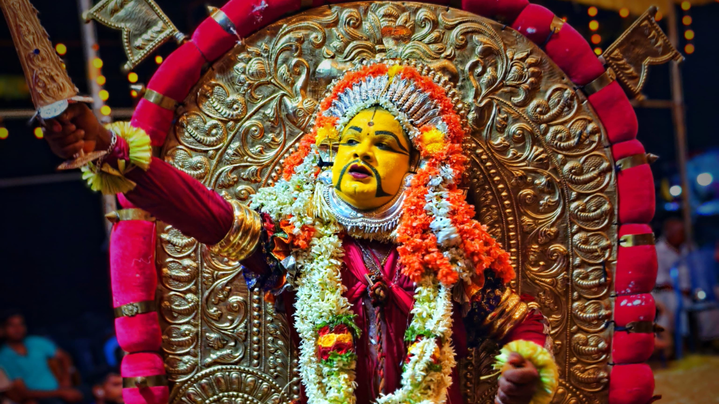 A Bhoota Kola performer in full regalia (yellow face paint, silver crown and flower garlands) dancing before a great embossed golden disc, brandishing a ceremonial sword at night
