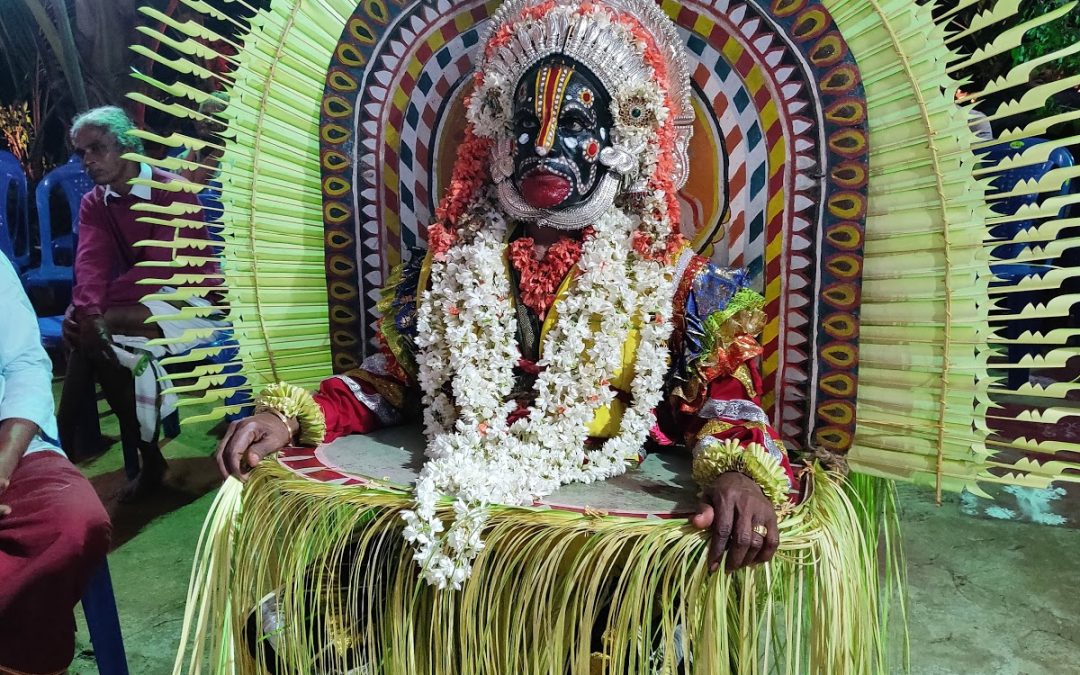 A Bhoota Kola performer with a black-and-white painted face, seated in state before a great fan of split tender coconut fronds, draped in white flower garlands while a villager attends