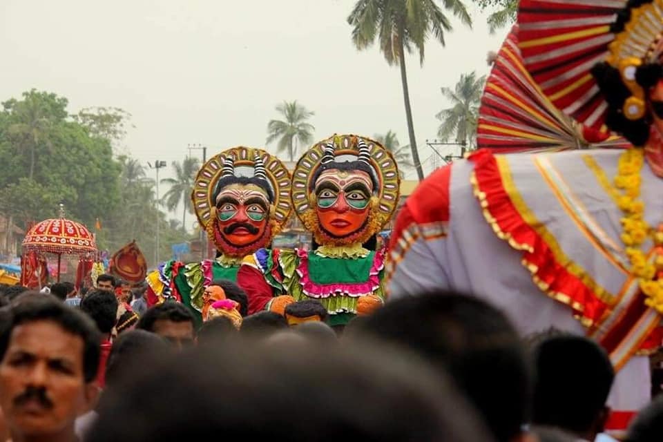 Two performers in towering painted masks and flower-ringed crowns move through a dense festival crowd during a Daiva procession in Kundapura