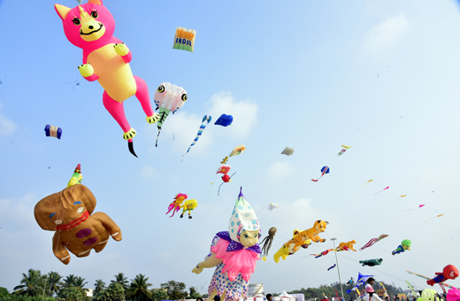 Large colourful animal-shaped kites and streamers fill a clear blue sky above a coastal beach during a kite festival