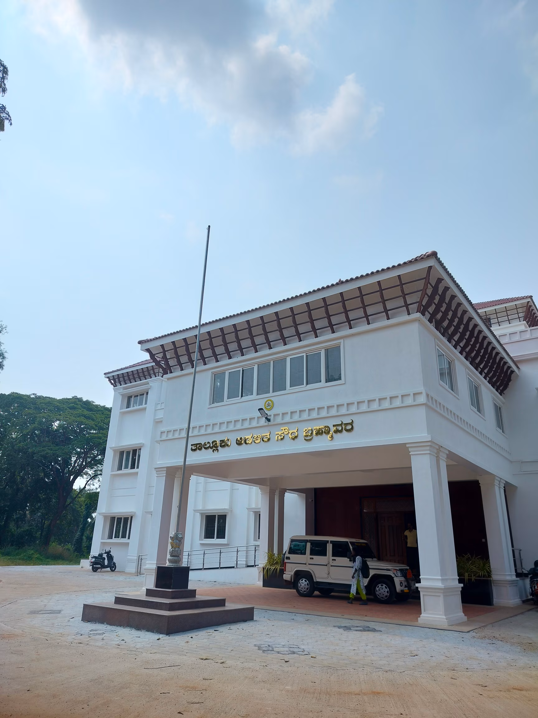 A modern taluk administrative office building (Taluka Soudha) in Udupi district, white with a tiled portico, flagpole and the Ashoka emblem