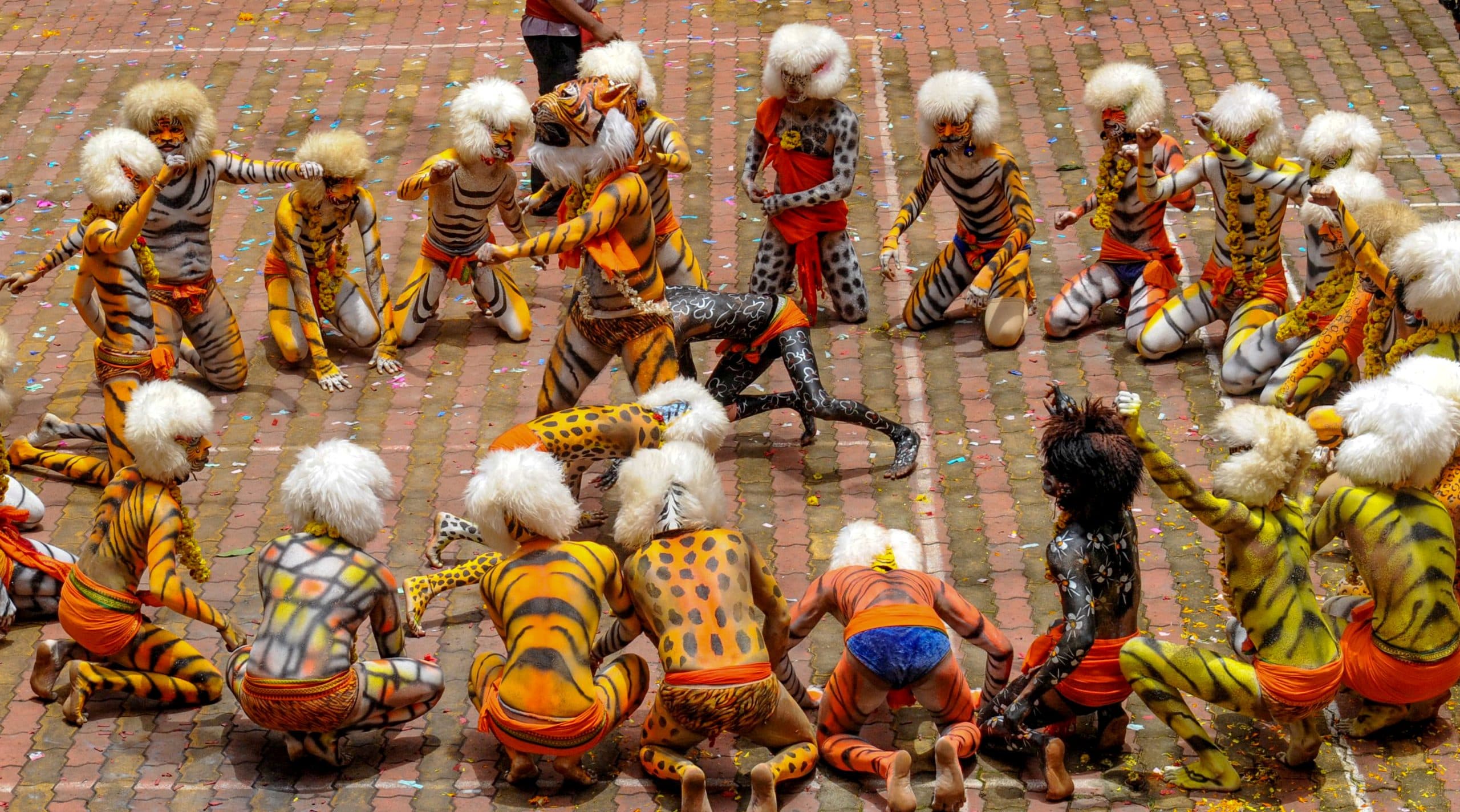 An overhead view of dozens of tiger-painted Huli Vesha dancers performing in a circle on a paved temple square scattered with confetti