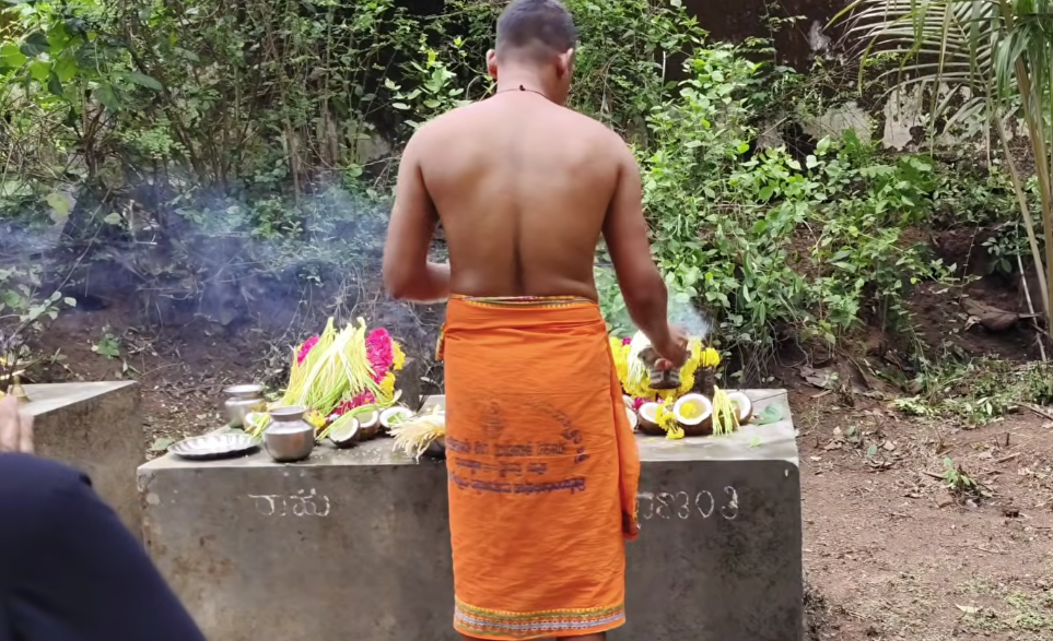 A priest in an orange dhoti performing Jakni puja at a roadside Daiva shrine, with coconut, flowers, brass vessels and rising incense smoke amid green foliage