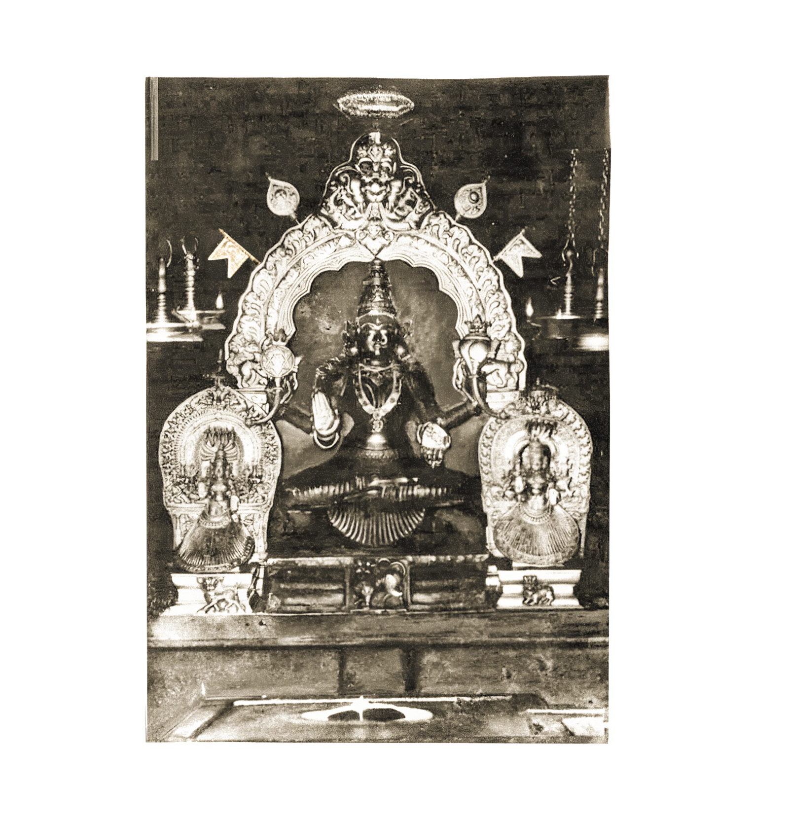 The oldest known photograph of Goddess Mookambika Devi at Kollur, a sepia image of the four-armed idol beneath its carved prabhavali arch, flanked by two attendant goddesses and ranks of brass oil lamps
