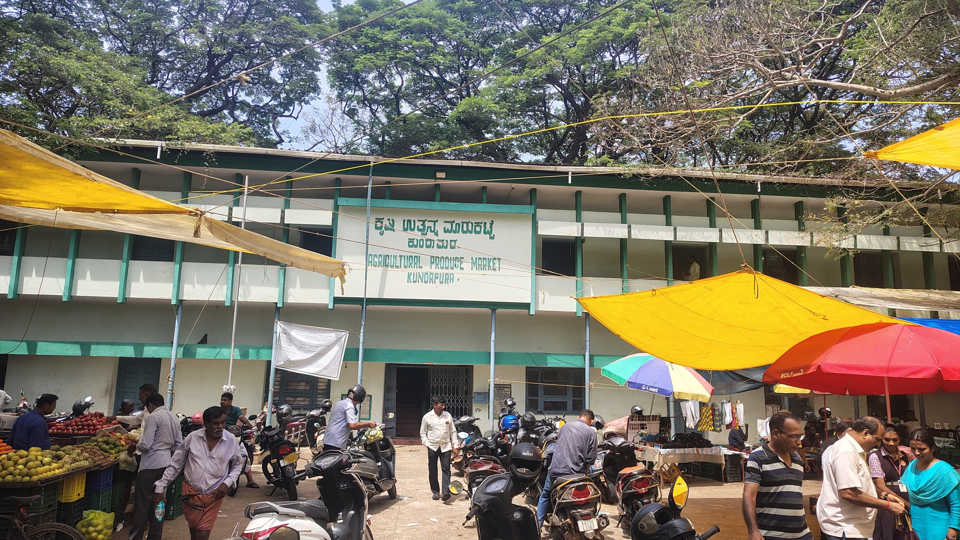 The Agricultural Produce Market (APMC) at Kundapura, fruit stalls and shoppers spilling out under bright tarpaulins and old rain trees