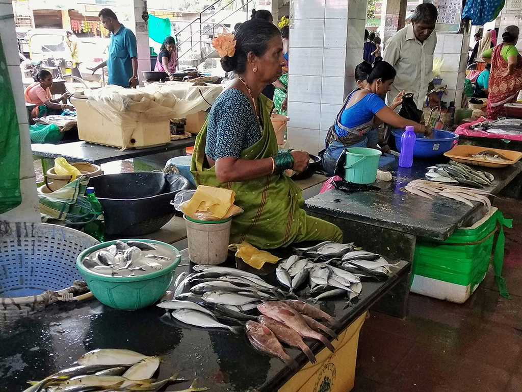 A vendor seated at a wet stone slab in the Kundapura fish market, fresh mackerel and sardines spread out before her with bowls and baskets around